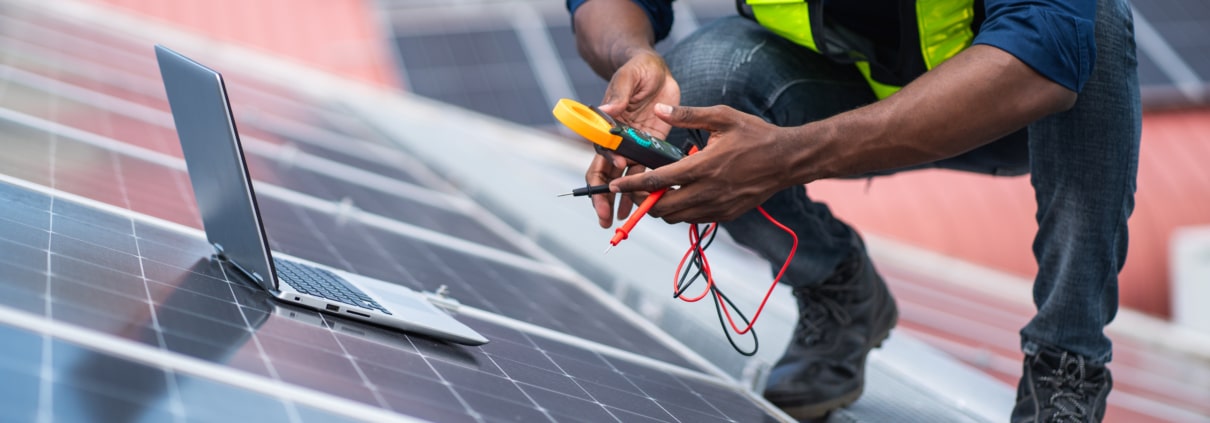 Technician testing rooftop solar panels with an electrical meter beside a laptop