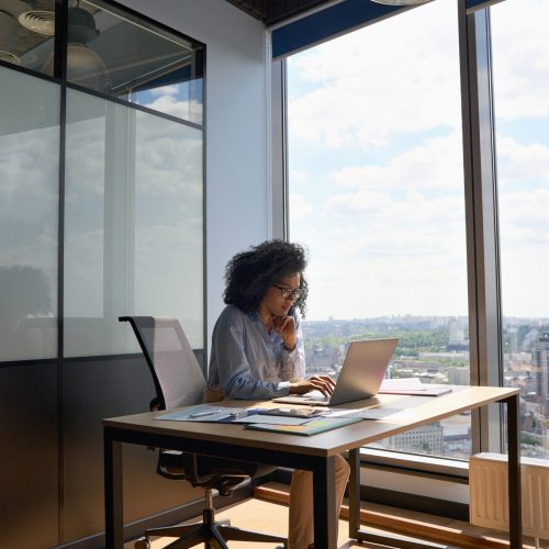 Concentrated African American executive top manager sitting at desk working typing on laptop computer in modern corporate office with panoramic view. Business technologies concept. Vertical shot.