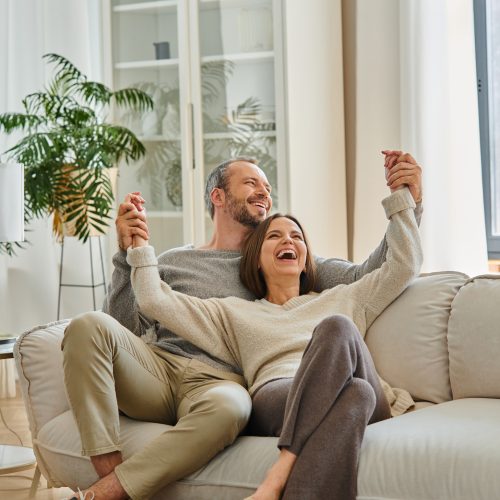 excited child-free couple holding hands and laughing on cozy couch in living room, fun and leisure